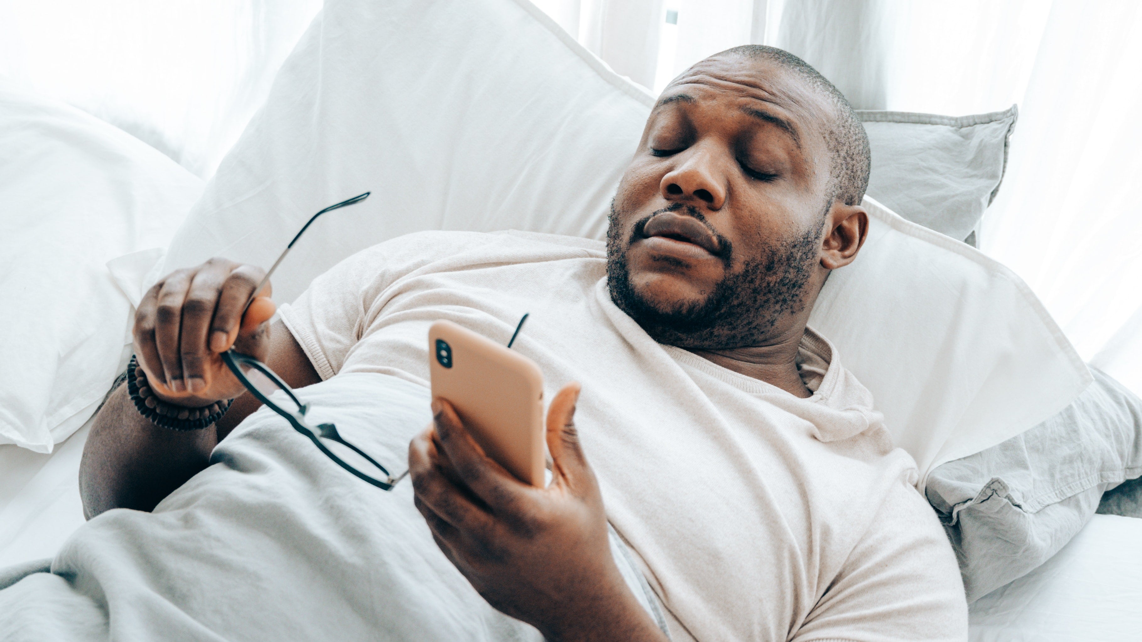 Homem negro deitado segurando óculos com uma mão e celular com outra